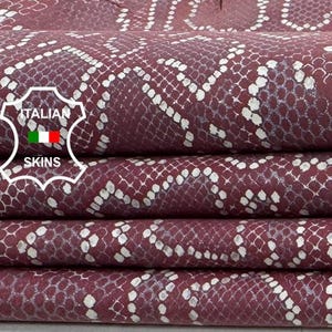 Peut inclure: Gros plan sur des feuilles de cuir bordeaux empilées avec un motif peau de serpent. Le cuir présente un motif argenté métallique et est plié avec soin. L'image comprend le logo Italian Skins.