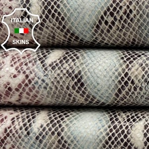 Op de afbeelding: Drie op elkaar gestapelde rollen slangenleer. Het leer heeft een patroon van lichtblauwe, witte en bordeauxrode vlekken, bedekt met een fijn, ruitvormig netpatroon. Het Italian Skins-logo is zichtbaar.