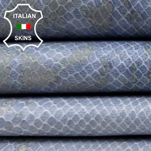 Puede incluir: Primer plano de hojas de cuero texturizadas apiladas en tonos azules y grises, con un patrón de piel de serpiente. El cuero tiene un brillo sutil y las capas están cuidadosamente dispuestas. El logotipo "Italian Skins" es visible.