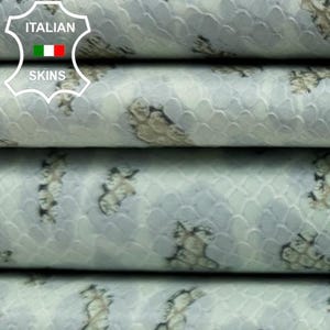 Può includere: Primo piano di fogli di pelle blu chiaro impilati con un motivo simile a un rettile in beige e nero. La pelle ha una lucentezza sottile e una superficie strutturata. Il logo della pelle italiana è visibile in alto a sinistra.