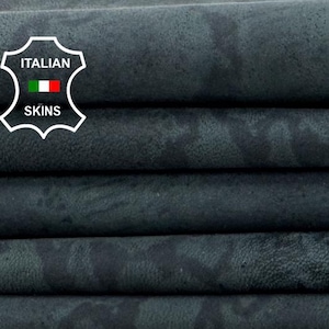 Puede incluir: Una pila de pieles de cuero gris oscuro con un patrón sutil y moteado. El cuero está doblado cuidadosamente, revelando la textura y las variaciones de color. El logotipo de Italian Skins es visible en la esquina superior izquierda.