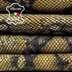 Op de afbeelding: Close-up van opgestapelde lederhuiden met een slangenpatroon. De huiden zijn een mix van goud en zwart, met een textuur, geschubd uiterlijk. Het Italiaanse lederlogo is zichtbaar in de linkerbovenhoek.
