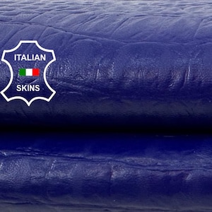 Könnte beinhalten: Nahaufnahme von zwei Stücken tiefblauen Leders mit einem strukturierten, krokodilartigen Muster. Das Leder ist gefaltet und gibt eine weiße Oberfläche darunter frei. Das Italian Skins-Logo ist in der oberen linken Ecke sichtbar.