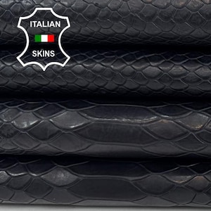 Puede incluir: Primer plano de pieles de cuero negras apiladas con textura de piel de serpiente. Las pieles están dobladas, revelando la superficie texturizada y la veta natural del cuero. La imagen incluye el logotipo "Italian Skins".