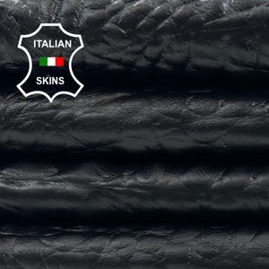 May include: Close-up of stacked black leather hides with a textured, embossed pattern. The leather appears to be folded over itself, revealing the layers. The Italian Skins logo is visible in the upper left corner.
