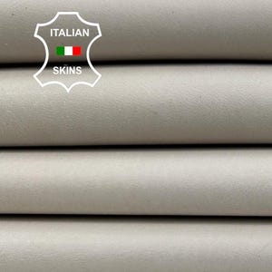 May include: A stack of light gray leather hides. The hides are neatly folded, revealing a smooth, matte texture. The image includes a logo with the text "Italian Skins".