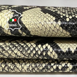 Op de afbeelding: Stapel van drie beige en zwarte slangenleerhuiden. De huiden zijn glad en hebben een glanzende afwerking. De huiden zijn op elkaar gestapeld, met de bovenste huid het meest zichtbaar. De huiden zijn gelabeld met "Italian Skins".