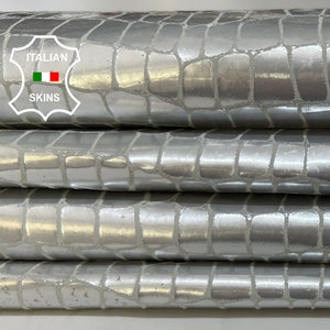 May include: Close-up of four rolls of silver-colored embossed leather. The leather has a crocodile pattern and is stacked on top of each other. The label "ITALIAN SKINS" is visible on the top roll.
