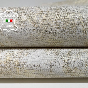 May include: Close-up of a white leather with a subtle gold embossed pattern. The leather is folded in thirds, showing the texture and sheen.