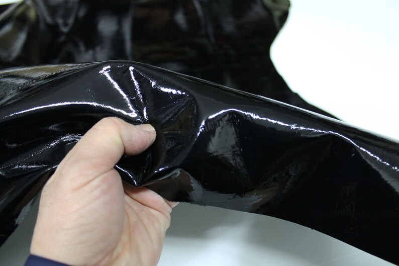 May include: Black patent leather with a glossy finish. The leather is folded and held in a hand.