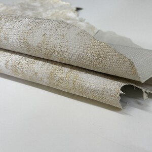 May include: Close-up of a folded piece of white leather with a subtle gold shimmer. The leather has a textured surface and a slightly distressed look. The label "ITALIAN SKINS" is visible in the top left corner.