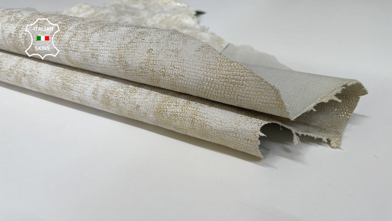 May include: Close-up of a folded piece of white leather with a subtle gold shimmer. The leather has a textured surface and a slightly distressed look. The label "ITALIAN SKINS" is visible in the top left corner.
