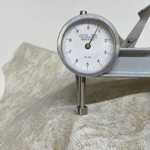 May include: A close-up of a leather sample being measured with a dial gauge. The gauge reads 5.7 mm. The leather is a light beige color with a subtle pattern.