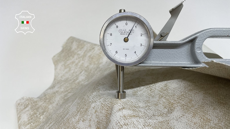 May include: A close-up of a leather sample being measured with a dial gauge. The gauge reads 5.7 mm. The leather is a light beige color with a subtle pattern.