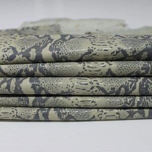 May include: Close-up of a stack of beige faux snakeskin fabric with a dark gray pattern. The fabric is folded and has a textured surface.