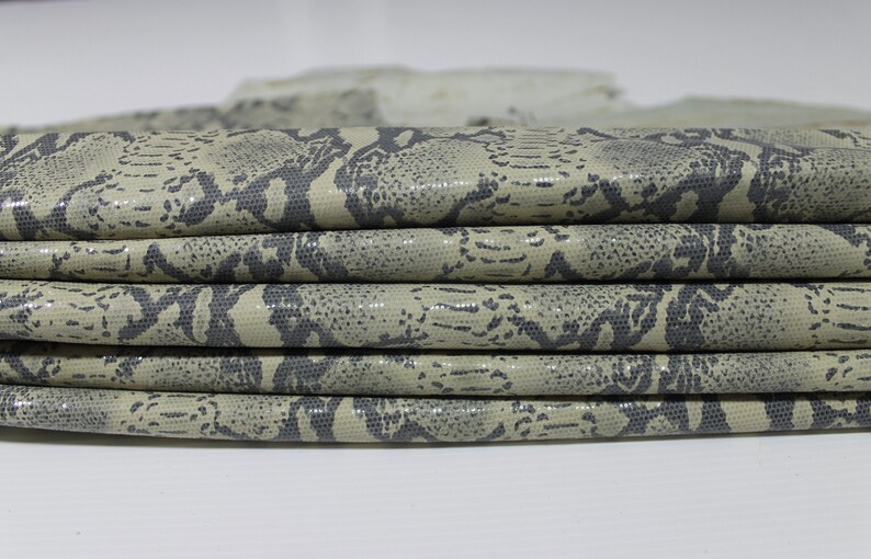 May include: Close-up of a stack of beige faux snakeskin fabric with a dark gray pattern. The fabric is folded and has a textured surface.