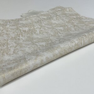 May include: A piece of off-white leather with a subtle gold flecked pattern. The leather is folded in half and has a rough, textured surface.