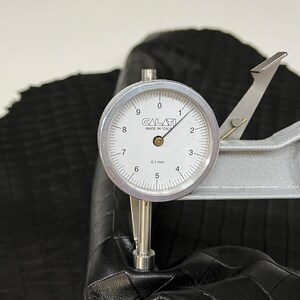 May include: A close-up of a leather sample being measured with a dial gauge. The gauge reads 3.1 mm. The leather is black and has a textured surface.