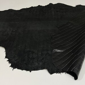 May include: Black leather hide with a textured surface. The leather is folded in half, revealing a smooth, patterned side.