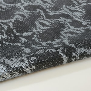May include: Close-up of a black and silver snakeskin leather fabric. The fabric has a textured, scaly pattern. The fabric is folded in half and the edges are visible. The fabric is labelled "ITALIAN SKINS".