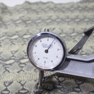 May include: A close-up of a leather thickness gauge with a dial reading 0.1 mm. The gauge is resting on a piece of green and white patterned fabric.