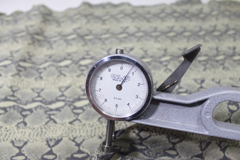 May include: A close-up of a leather thickness gauge with a dial reading 0.1 mm. The gauge is resting on a piece of green and white patterned fabric.
