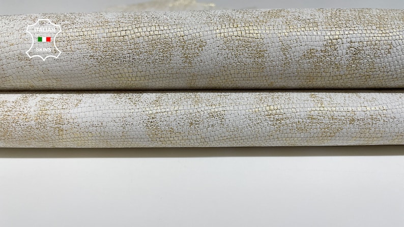 May include: Close-up of a white leather with a subtle gold embossed pattern. The leather is folded in thirds, showing the texture and sheen.