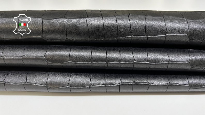 May include: Close-up of three black crocodile embossed leather sheets stacked on top of each other. The leather has a subtle sheen and a textured surface. The label "ITALIAN SKINS" is visible in the top left corner.