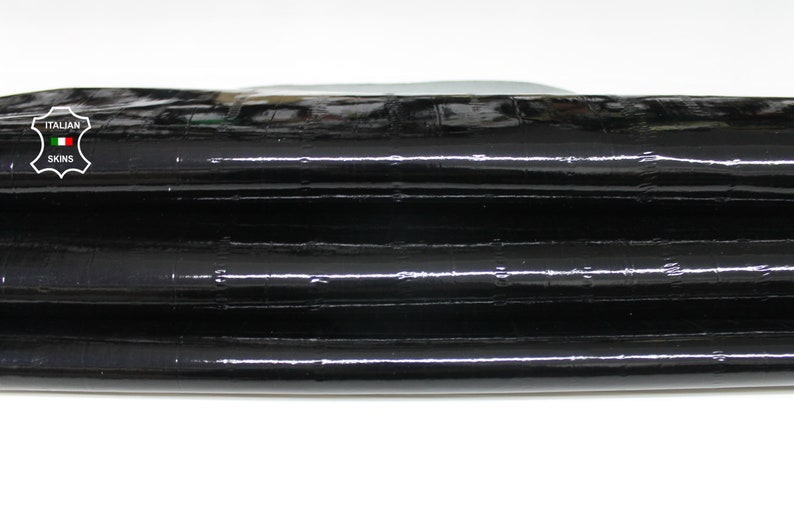 May include: Close-up of black leather with a glossy finish. The leather is folded in thirds, revealing the texture of the material. The leather is labeled "ITALIAN SKINS".