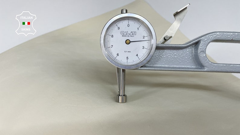 K&ouml;nnte beinhalten: Nahaufnahme einer Lederprobe, die mit einem Messger&auml;t gemessen wird. Das Messger&auml;t zeigt 0,1 mm an und wurde in Italien hergestellt. Das Leder ist hellbeige.