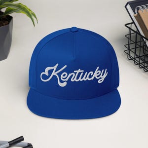 Op de afbeelding: Een koningsblauwe baseballcap met het geborduurde woord "Kentucky" in wit script. De cap heeft een platte klep en een gestructureerde kroon. De hoed wordt gepresenteerd op een wit oppervlak.