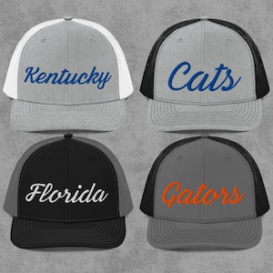 Puede incluir: Cuatro gorras de camionero con la parte trasera de malla. Las gorras son grises, negras y blancas, con las palabras "Kentucky", "Cats", "Florida" y "Gators" bordadas en la parte delantera en azul, blanco y naranja.