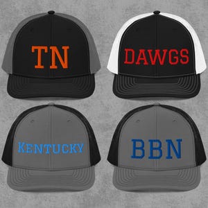 Puede incluir: Cuatro gorras de camionero con diferentes combinaciones de colores. La primera es negra con la parte trasera de malla gris y las letras "TN" en naranja. La segunda es negra con la parte trasera de malla blanca y la palabra "DAWGS" en rojo. La tercera es gris con la parte trasera de malla negra y la palabra "KENTUCKY" en azul. La cuarta es gris con la parte trasera de malla negra y las letras "BBN" en azul.