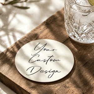 以下が含まれることがあります： 中央に黒い筆記体で「Your Custom Design」と印刷された、オフホワイトの円形コースター。コースターは滑らかな表面をしており、氷とライムのスライスが入った透明なグラスの横に置かれています。