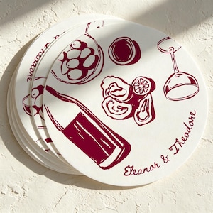 Pode incluir: Uma pilha de bases para copos redondas, de cor branco-sujo, com uma ilustração de linhas vermelho-escuras de uma garrafa de vinho, um copo, uma taça de azeitonas e ostras. O texto "Eleanor & Theodore" está impresso em cursivo na base.