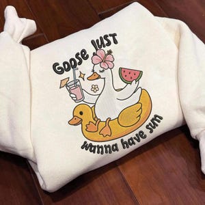 Peut inclure: Sweat-shirt crème avec un dessin d'oie portant une fleur et tenant une boisson et une pastèque. L'oie est sur un flotteur en forme de canard jaune. Le texte dit "Goose Just Wanna Have Sum".