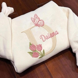 Peut inclure: Sweat-shirt crème avec broderie. Il présente une grande lettre "D" beige, ornée d'un papillon rose, de tulipes roses et du nom "Daiana" en écriture rose. Le sweat-shirt a des poignets côtelés et un col rond.