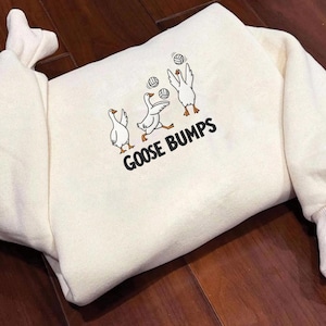 May include: Cream-colored sweatshirt with a playful graphic of three cartoon geese playing volleyball. The text "GOOSE BUMPS" is printed in black below the geese. The sweatshirt has ribbed cuffs and a crew neck.