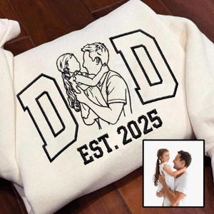Puede incluir: Sudadera de color crema con un diseño bordado en negro. El diseño presenta la palabra "DAD" en letras negritas, un dibujo lineal de una persona sosteniendo a un niño y el texto "EST. 2025". Una pequeña foto enmarcada está en la esquina inferior derecha.