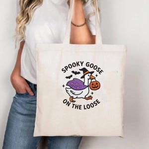 May include: A cream-colored canvas tote bag featuring an embroidered graphic of a white goose wearing a black witch hat and a purple cape with orange stars. The goose holds an orange jack-o'-lantern, surrounded by black bats and the text SPOOKY GOOSE ON THE LOOSE.