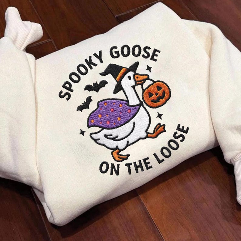 May include: A cream-colored sweatshirt featuring an embroidered graphic of a white goose wearing a black witch hat and a purple polka-dot cape. The goose holds an orange jack-o'-lantern bucket, with black bats and the text SPOOKY GOOSE ON THE LOOSE.