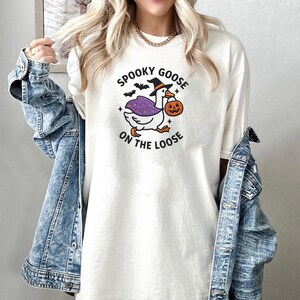 May include: A white t-shirt featuring a graphic of a goose wearing a witch hat and a purple cape with orange stars, carrying a jack-o'-lantern. The text reads SPOOKY GOOSE ON THE LOOSE, accompanied by small black bats and stars. A blue denim jacket is worn over the shirt.