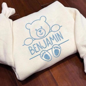 May include: A cream-colored sweatshirt featuring light blue embroidery of a teddy bear head and paws. The design includes the name BENJAMIN embroidered in light blue capital letters between the bear's head and its two feet, all outlined in blue.