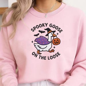 May include: A light pink crewneck sweatshirt featuring an embroidered graphic of a white goose wearing a witch hat and a purple cape with orange dots. The goose carries an orange jack-o'-lantern bucket. Text reads: SPOOKY GOOSE ON THE LOOSE.