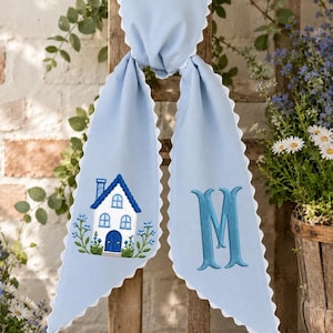 May include: A light blue fabric scarf with white scalloped edges. The left side features an embroidered white house with a blue roof and blue flowers. The right side features a large, embroidered blue letter M in a serif font style.