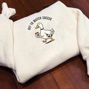 May include: Cream-colored sweatshirt with the text "OFF TO WATCH SOCCER" and a cartoon duck graphic. The duck is holding a coffee cup and a soccer ball, with a black outline and yellow beak and feet.