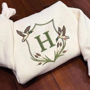以下が含まれることがあります： クリーム色のスウェットシャツ。刺繍された紋章には、2羽の飛んでいるアヒル、文字「H」、緑色のガマが描かれています。スウェットシャツはクルーネックとリブ編みの袖口が付いています。