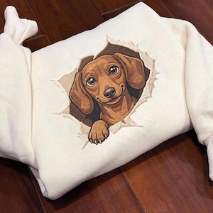 Puede incluir: Una sudadera de color crema con un bordado detallado de un perro salchicha marrón que parece asomarse a través de un agujero rasgado en la tela. El perro tiene ojos grandes, orejas caídas y una pata visible, representado en tonos marrones.