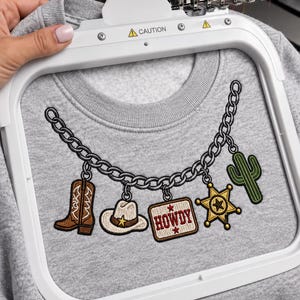 May include: A heather grey sweatshirt featuring an embroidered design of a silver chain necklace with five charms: a brown cowboy boot, a white cowboy hat, a rectangular sign with the text HOWDY, a yellow sheriff star, and a green cactus.