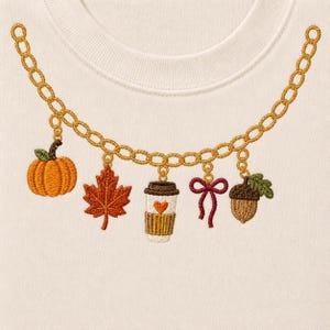 May include: A cream-colored sweatshirt featuring an embroidered gold chain necklace design. The chain is adorned with five charms: an orange pumpkin, a red maple leaf, a coffee cup with a heart, a maroon ribbon bow, and a brown acorn.